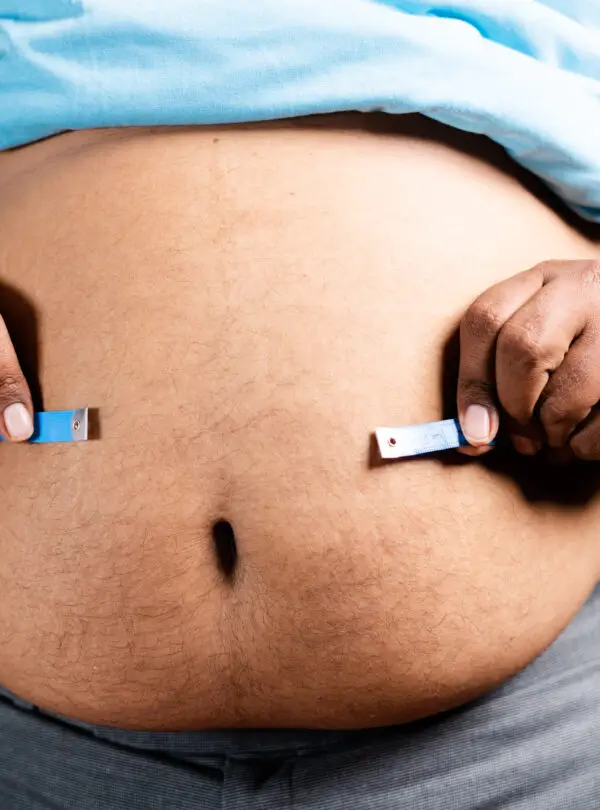 Unrecognizable unhealthy obese man checking stomach or waist fat using measuring tape before diet at home – concept of suffering body overweight due to junk food habits